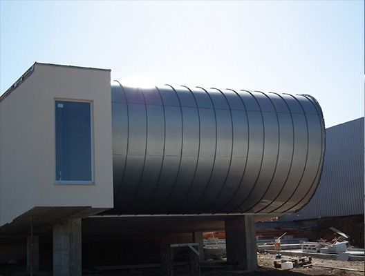 Edificio moderno con gran estructura metálica curva sobre pilares de hormigón, una sección en forma de caja blanca tiene una ventana.