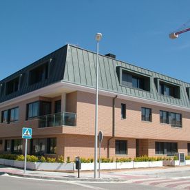 Edificio de ladrillo de tres pisos con techo de metal oscuro, acera y cartel de calle bajo un cielo azul.