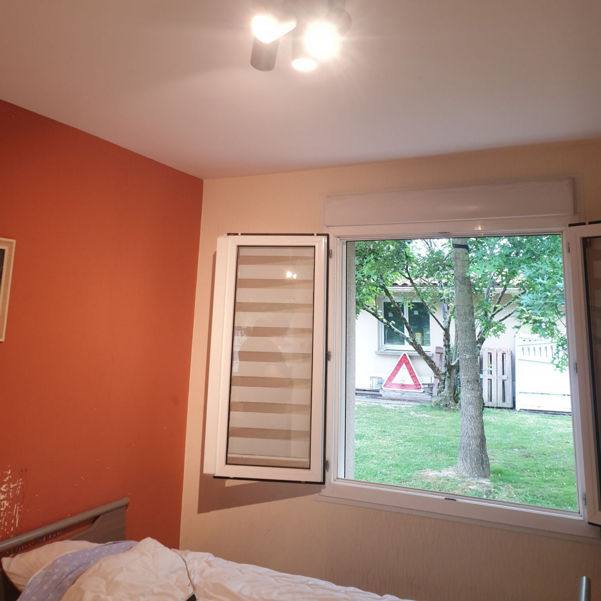 Chambre avec fenêtre ouverte donnant sur un jardin verdoyant et un arbre. Mur orange à gauche, beige à droite.