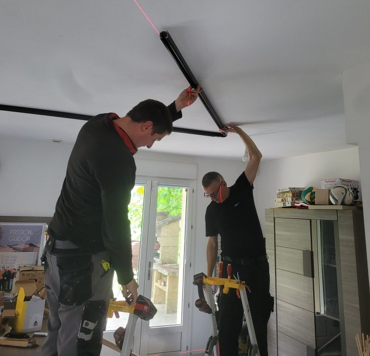 Deux personnes installent des poutres de plafond noires dans une pièce, à l'aide d'un niveau laser.