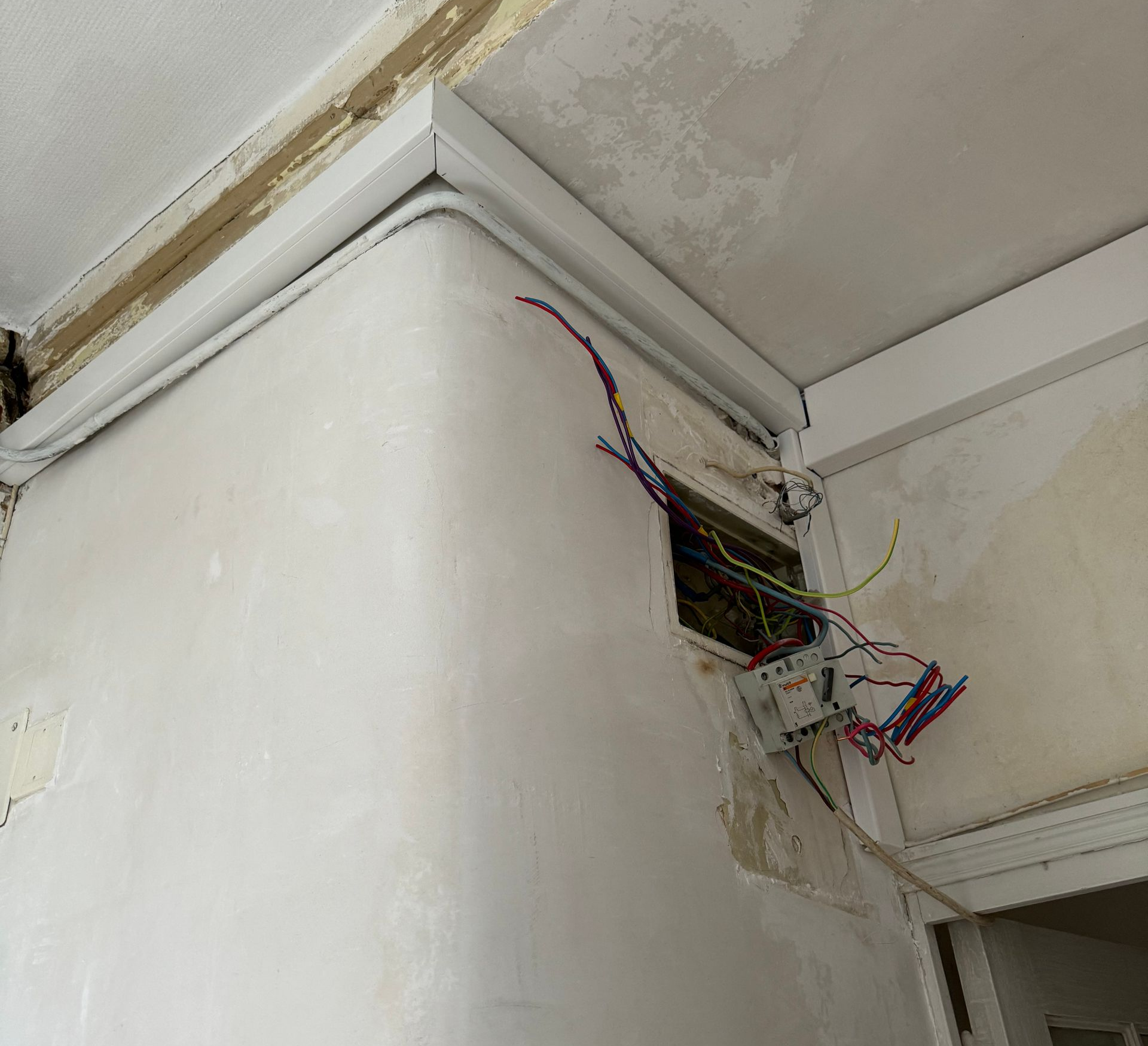 Des fils et des composants électriques apparents dépassent d'un mur endommagé, longeant un rail blanc près du plafond.