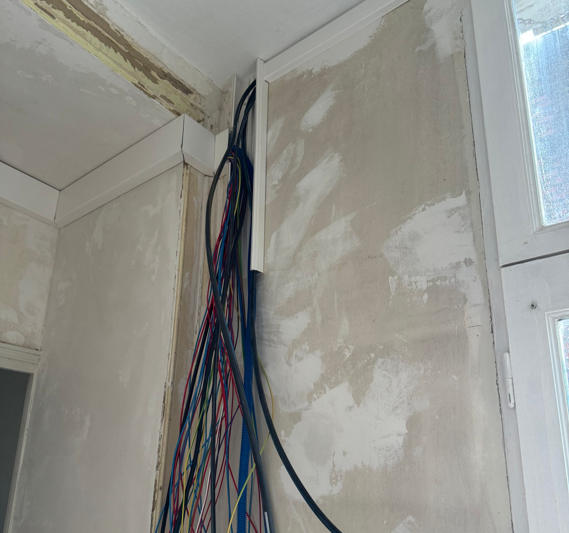 Des fils électriques courent verticalement le long d'un angle de mur, à l'intérieur d'une gaine blanche.