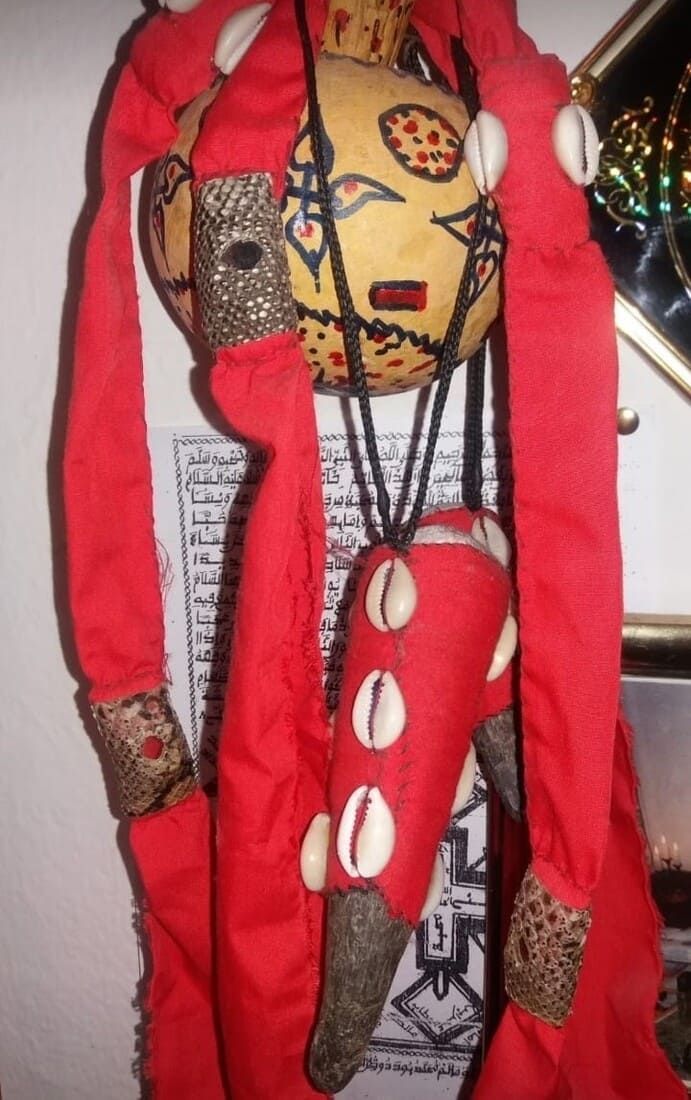Muñeca artesanal vestida de rojo, con largas trenzas y adornos de conchas, colgada frente a un fondo estampado
