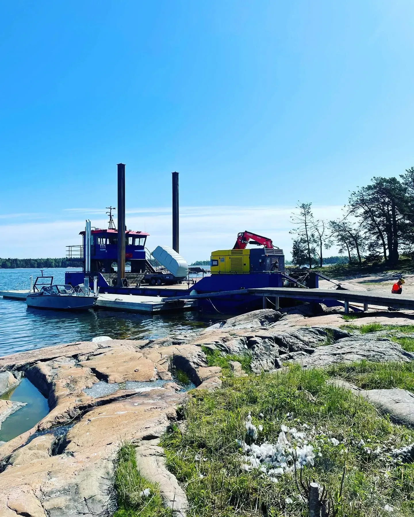 Sininen lautta telakoituna kallioiselle rannalle kirkkaan taivaan alla, kannella ajoneuvoja ja metallirampit näkyvissä.