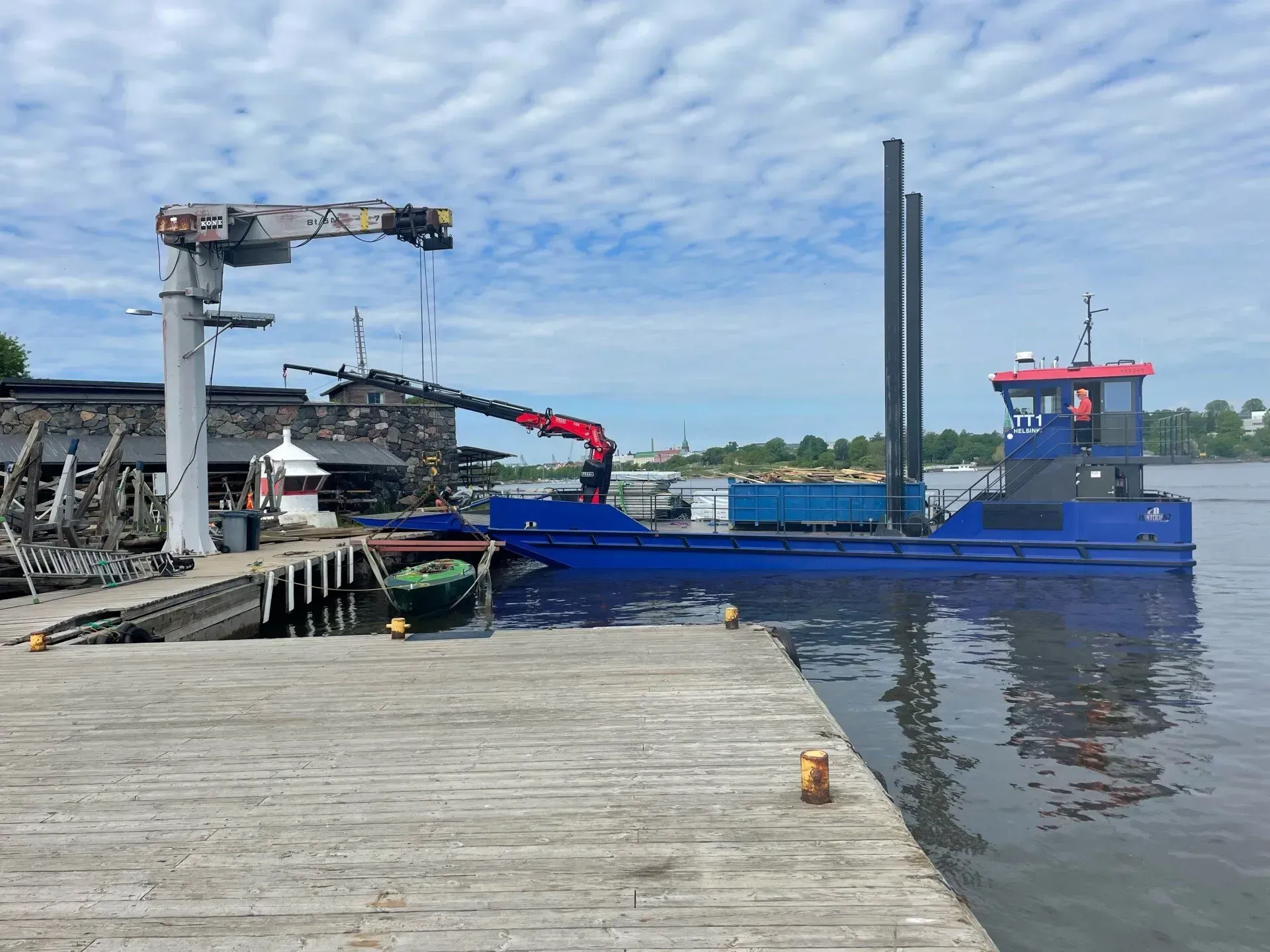 Sininen työvene telakoituna betonilaiturin viereen pilvisen taivaan alla, nostureita ja teollisuuslaitteita.