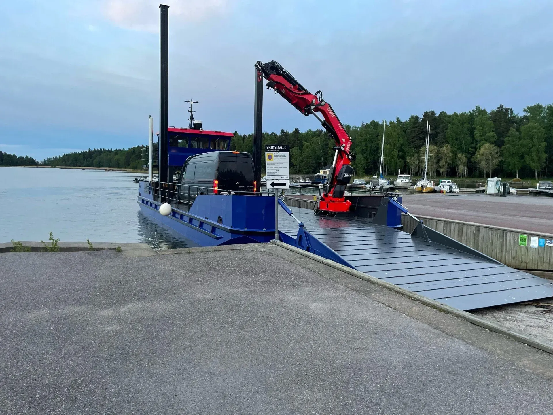 Punainen nosturi lastaa sinistä proomua rampilla varustetulla järvenrantalaiturilla.