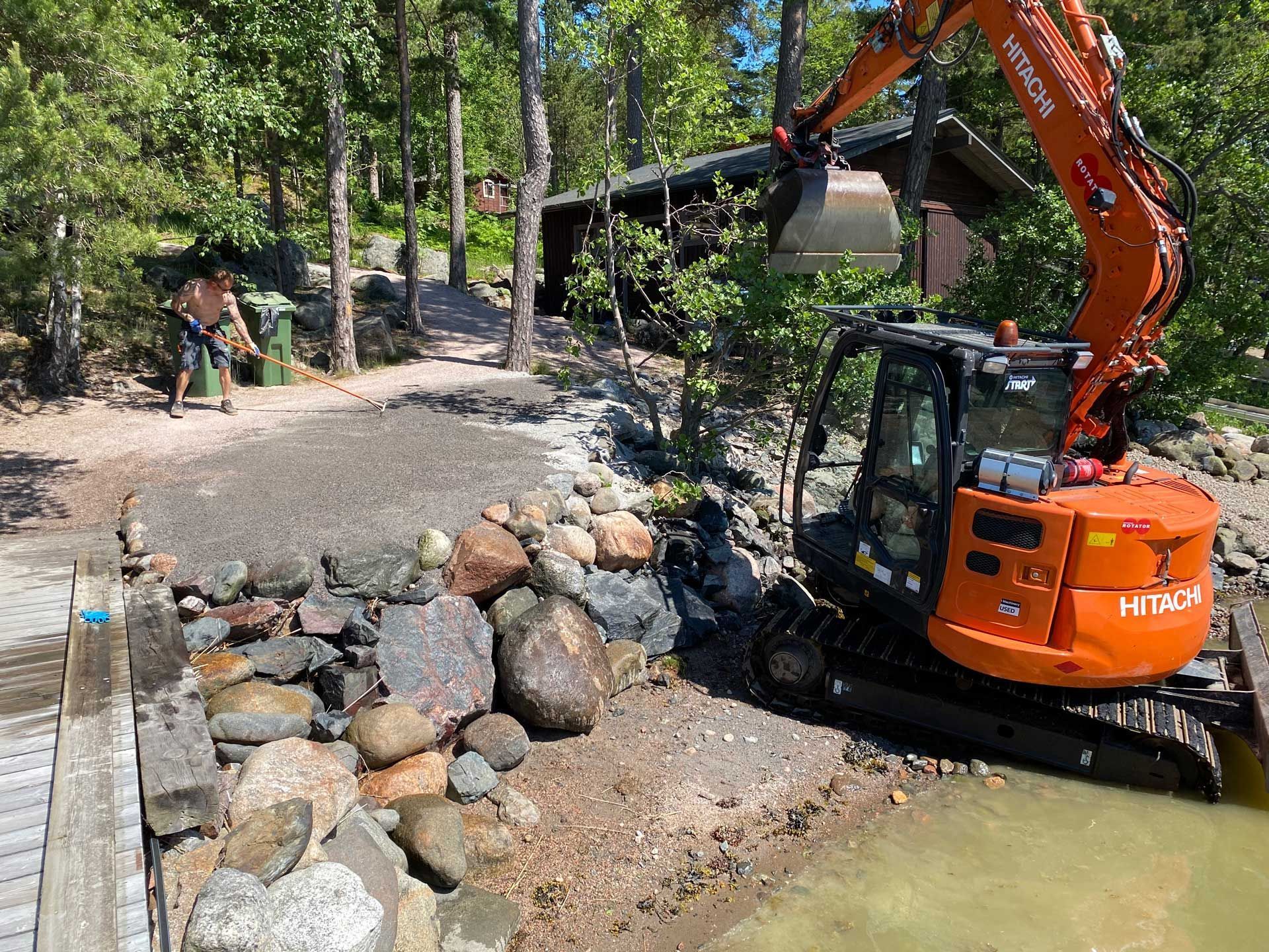 Oranssi kaivinkone kivisen puron vieressä, työntekijöitä ja hytti metsäisellä rakennusalueella.