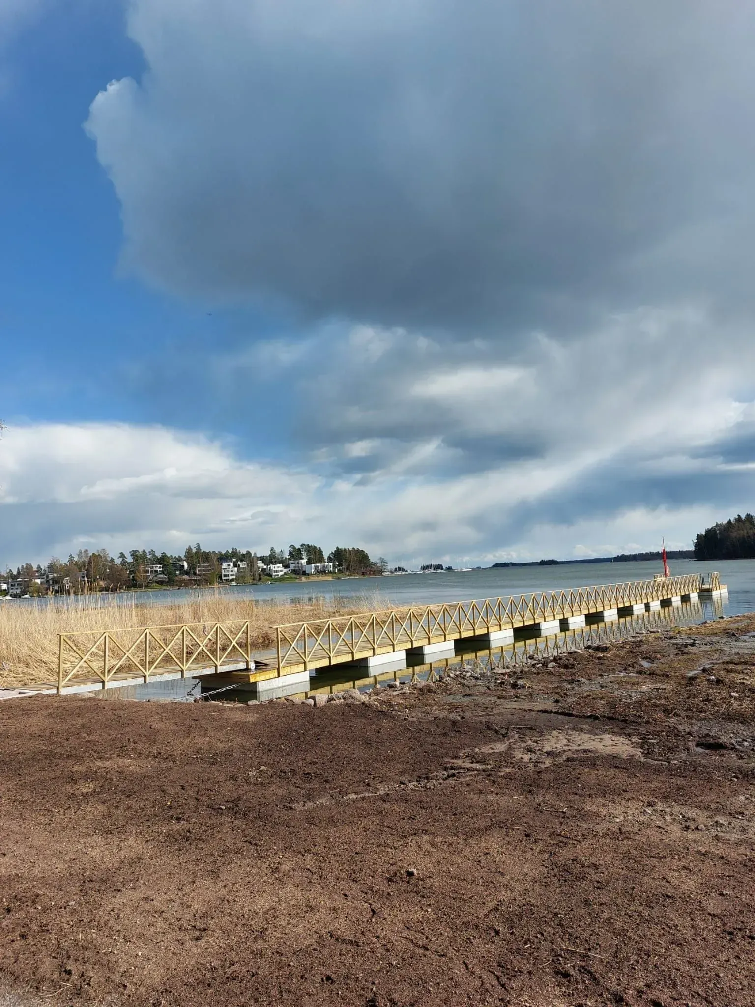 Pilvinen ranta, jossa on kiviä, merimuuri ja kaukaisia taloja rantaviivaa pitkin