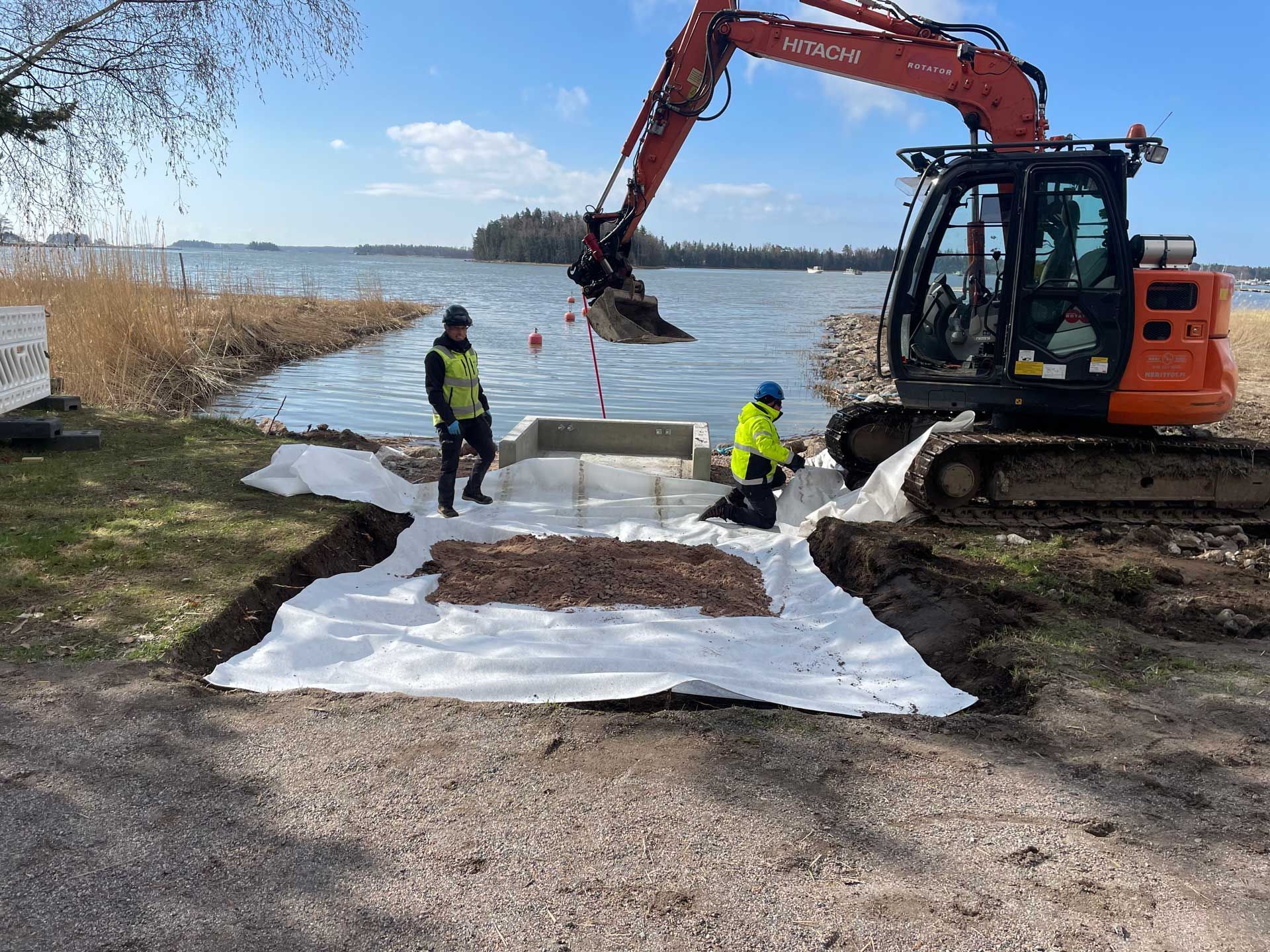 Kaivinkone ja työntekijät asentavat rantaviivaa järven rannalle.