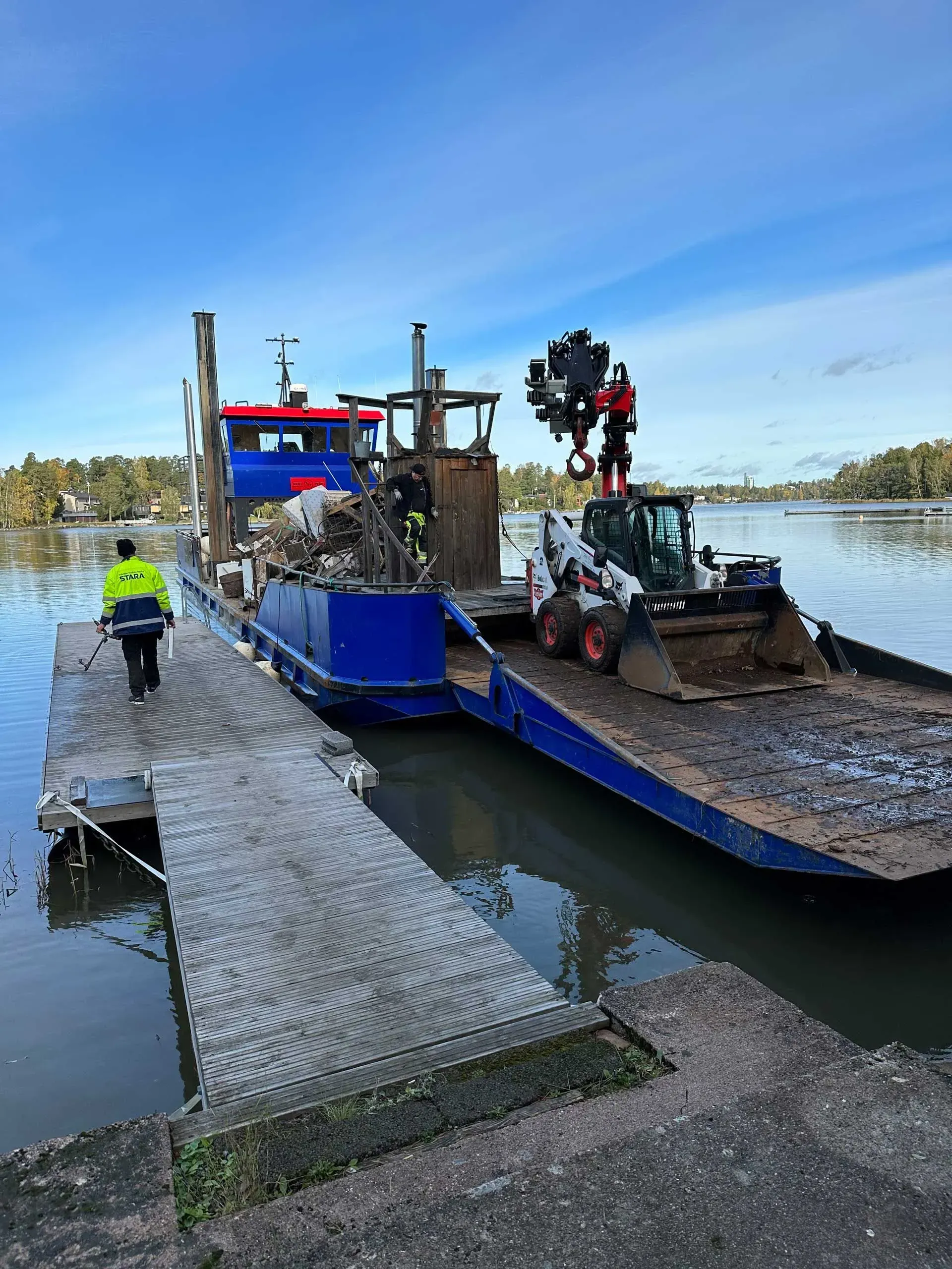 Sininen työproomu purkaa mutaista sedimenttiä rantalaiturilla, ja lähistöllä on työntekijä turvaliiveihin pukeutuneena.