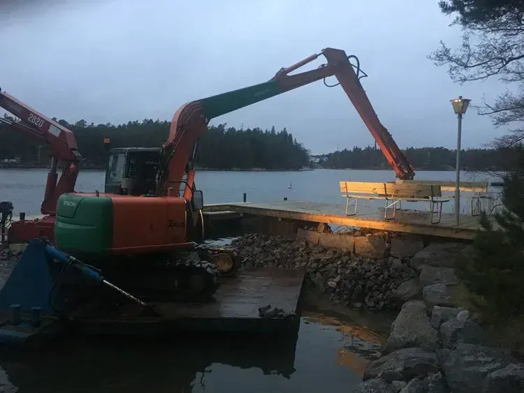 Kaivinkone lastaa maata proomuun joen varrella hämärässä, toisen kaivinkoneen ollessa lähistöllä.