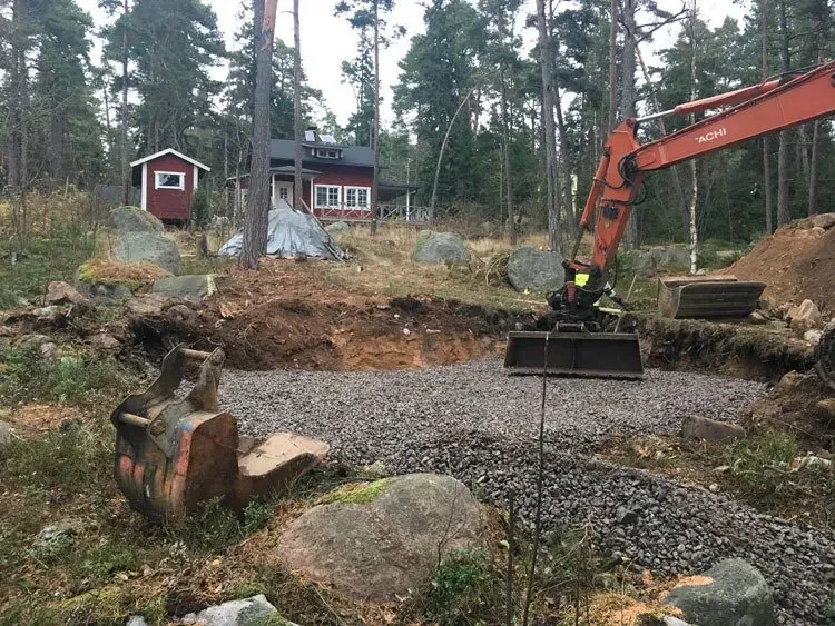 Kaivinkone kaivaa metsäisen mökkialueen lähellä, jossa on soraa, kiviä ja punainen talo taustalla