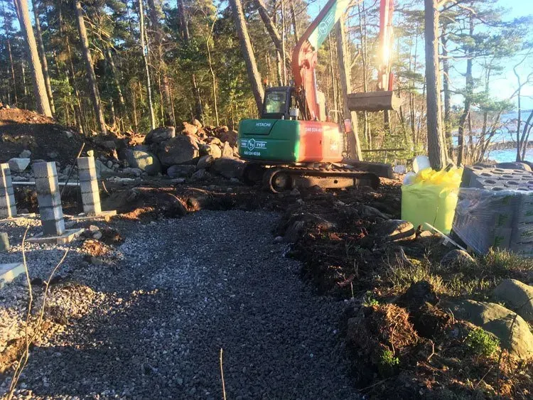 Pieni kaivinkone sorapolulla metsäisellä alueella järven rannalla, materiaali- ja roskapinojen kanssa.