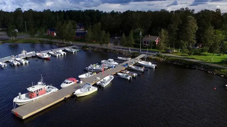 Pieni venesatama, jossa valkoiset veneet telakoituna tummaan veteen, metsän ja rantaviivan reunustama.