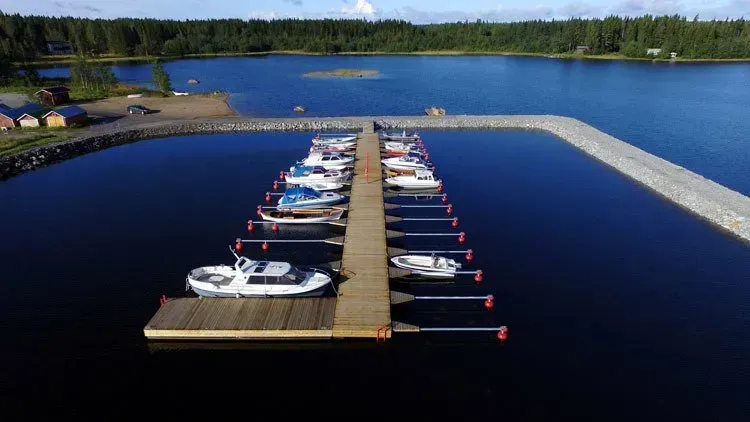 Venesatama, jonka veneet ovat telakoituna puisella laiturilla tyynessä tummansinisessä vedessä, kivisen aallonmurtajan ja metsän ympäröimänä.
