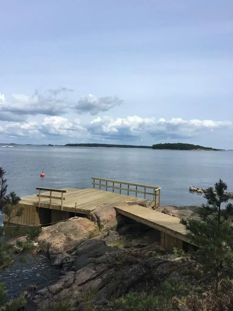 Puinen laituri kaiteilla kallioisella järvenrannalla pilvisen taivaan alla, tyynen veden ja kaukaisen maan takana.