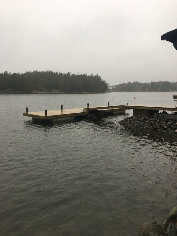 Sateinen järvenrantalaituri, jossa tyyni vesi, kivinen ranta ja taustalla puiden reunustama penger