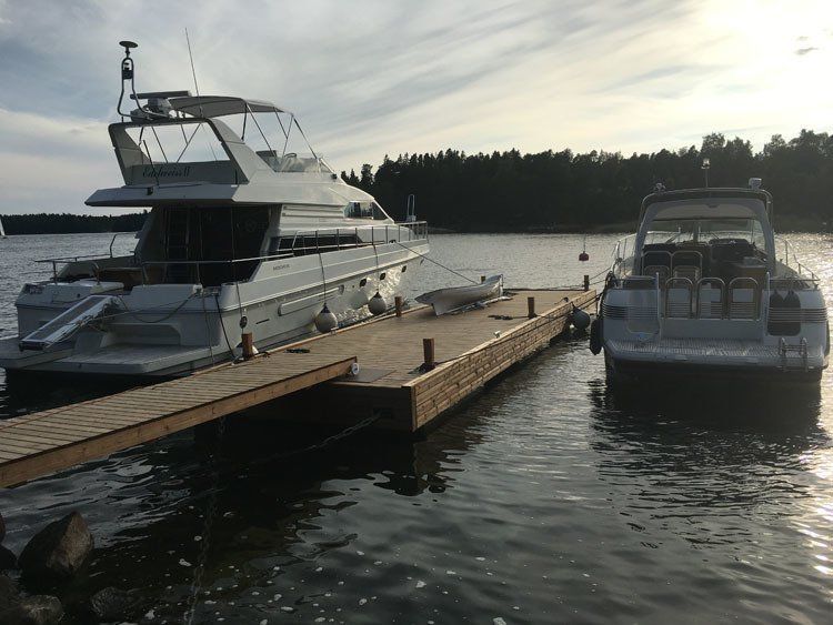 Kaksi jahtia telakoituna puiseen laituriin tyynellä vedellä lähellä puiden reunustamaa rantaa.