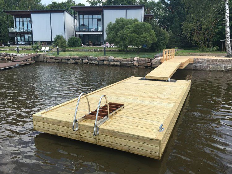 Järveen ulottuva puinen kelluva laituri, jonka takana rannalla on taloja ja puita.