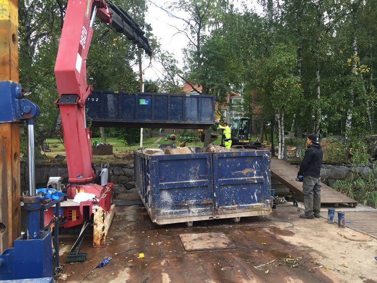 Rakennustyömaa, jossa punainen nosturi nostaa sinistä roskalavaa lähellä työntekijöitä metsäalueella