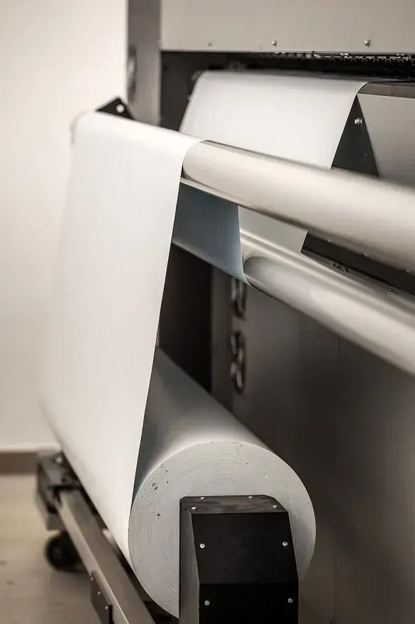 Impresora de gran formato con un rollo de papel blanco que sobresale. La máquina es gris y negra, colocada contra una pared blanca.