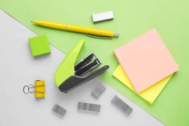 Una grapadora, un bolígrafo, notas adhesivas y clips para carpetas sobre un fondo verde y blanco.
