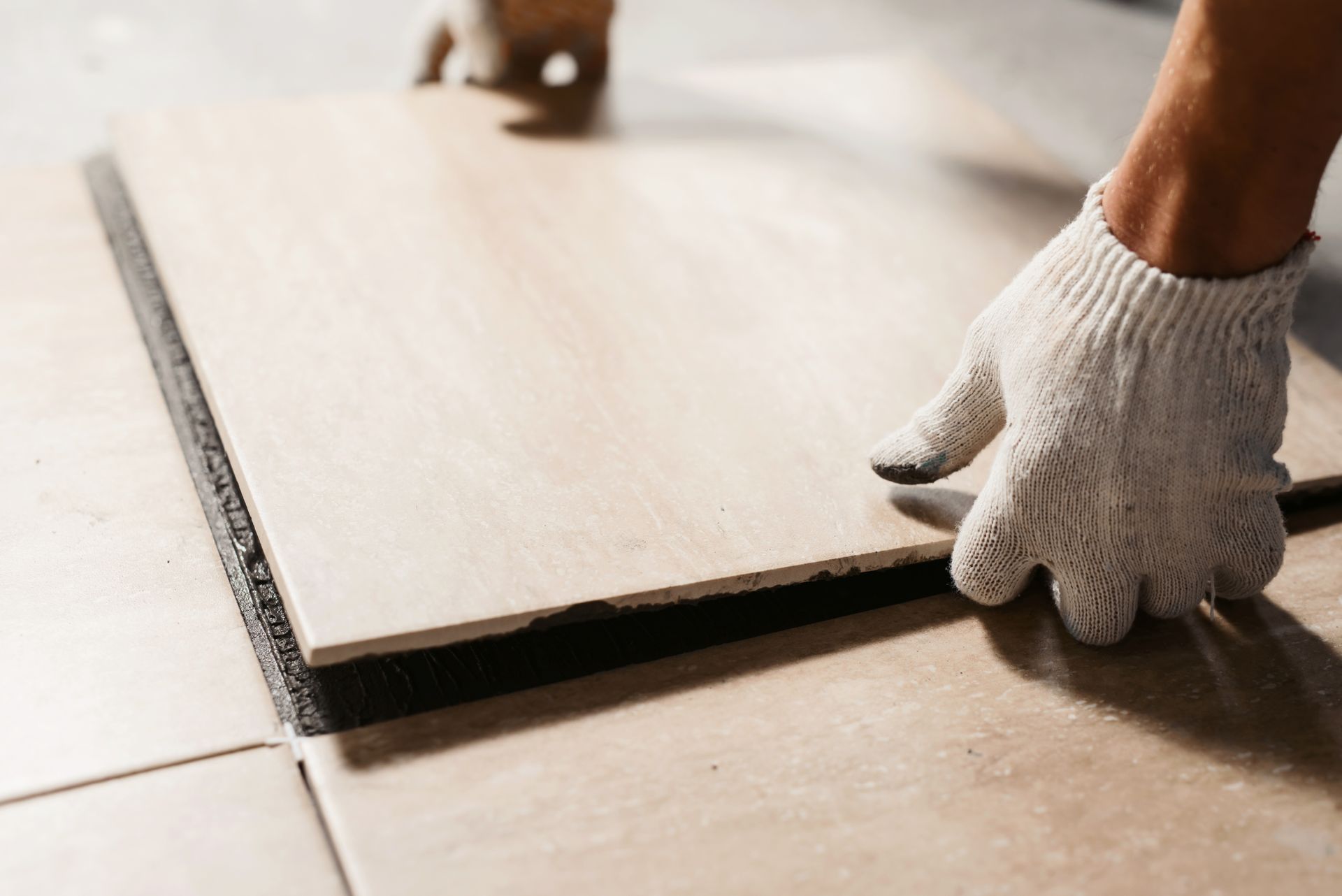Person mit Handschuhen, die eine beige Bodenfliese mit schwarzem Klebstoff auf einer Oberfläche verlegt.