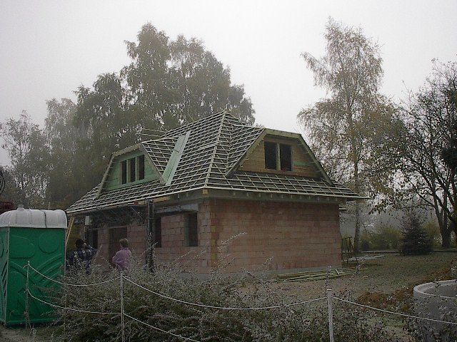 Haus im Bau mit freiliegenden Dachbalken, Fenstern und einer Ziegelwand. Grüne mobile Toilette im Vordergrund.