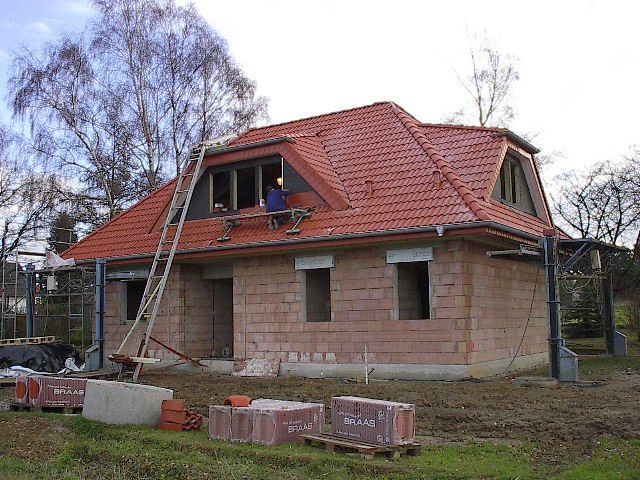 Haus im Bau: rotes Ziegeldach, Ziegelwände, Leiter, Person auf dem Dach, Materialien auf dem Boden.