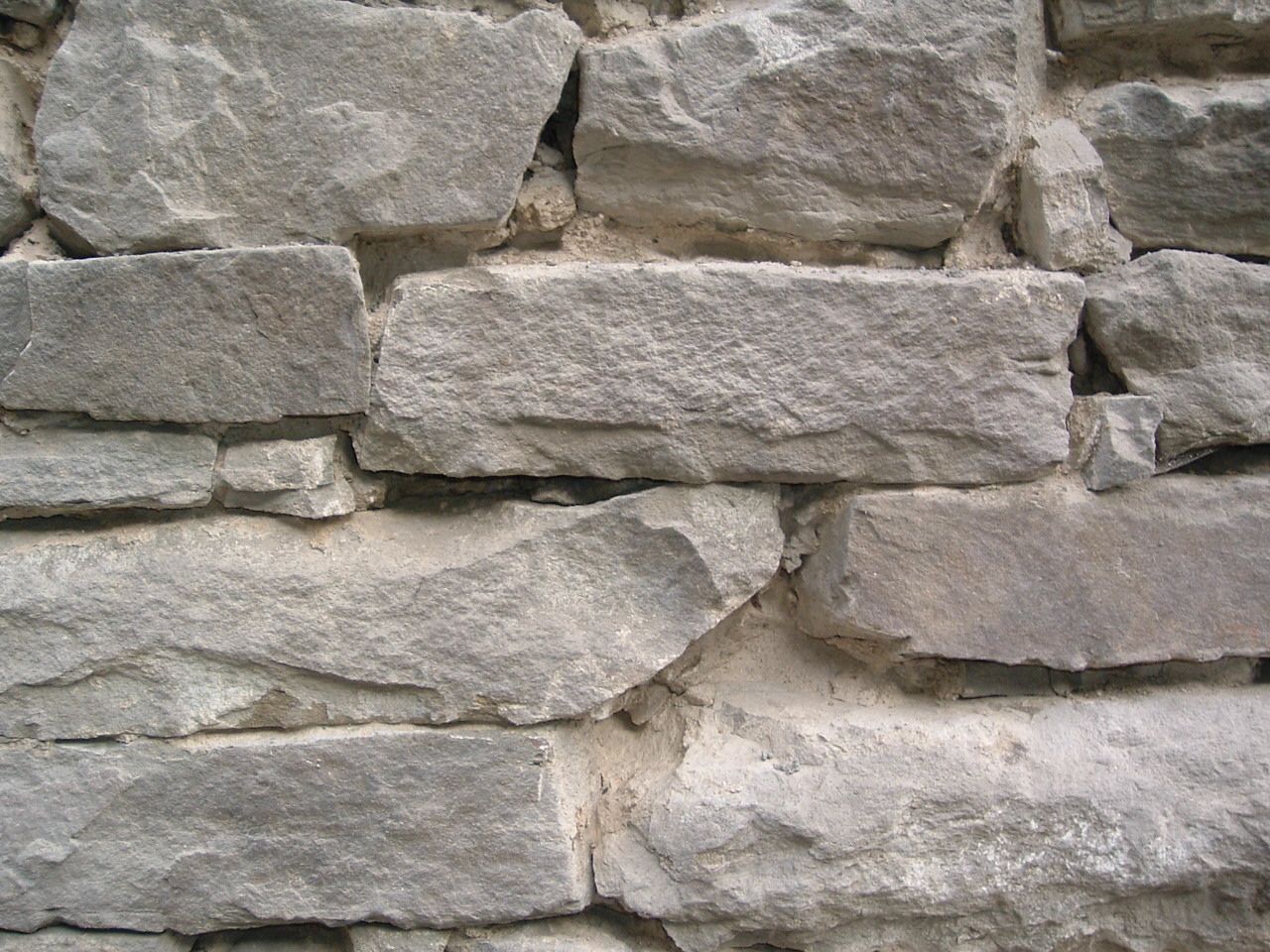 Nahaufnahme einer grauen Steinmauer mit rechteckigen und unregelmäßig geformten Steinen und Mörtel unterschiedlicher Größe.