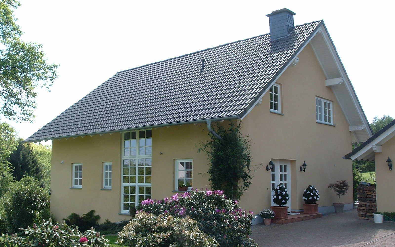 Gelbes Haus mit dunklem Ziegeldach, Haustür und Mehrscheibenfenstern; davor Rhododendren.