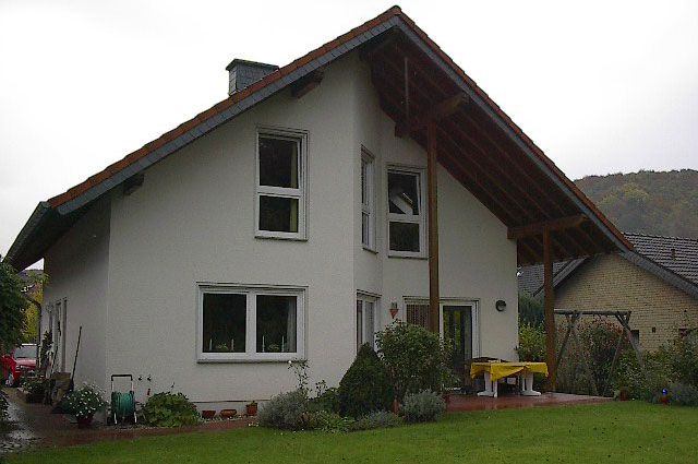 Zweistöckiges weißes Haus mit braunem Dach und Holzveranda, grünem Rasen, Fenstern und Pflanzen.