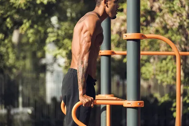Un hombre sin camisa está haciendo fondos en barras paralelas en un parque.
