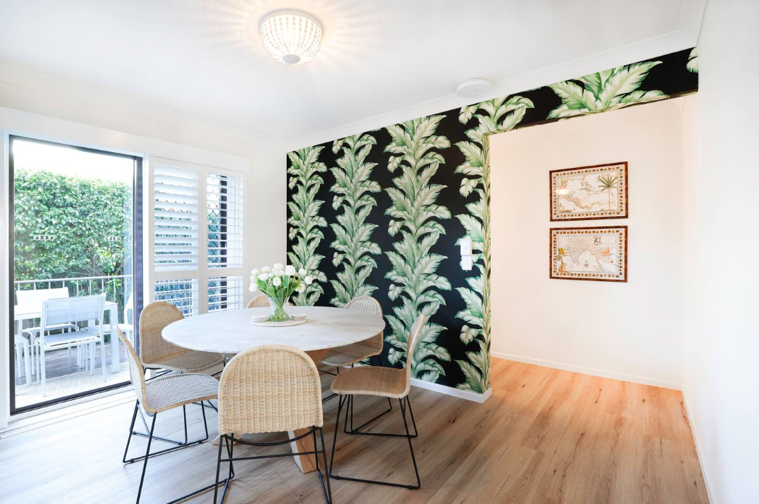 A dining room with a table and chairs and a tropical wallpaper on the wall.