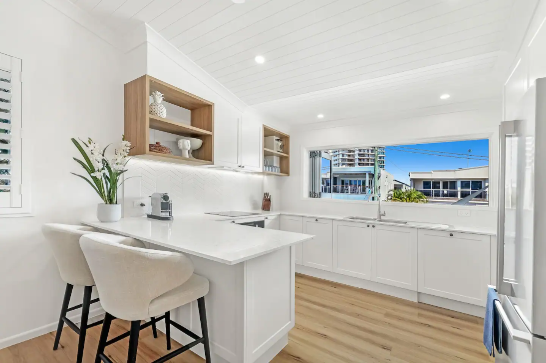 A kitchen with white cabinets , wooden floors , stools and a window.