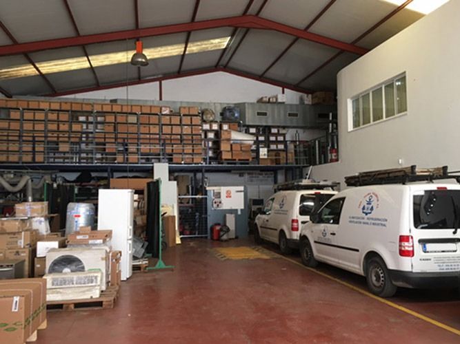 Interior de un almacén con cajas en estanterías y dos furgonetas aparcadas cerca. Suelo rojo, paredes blancas.