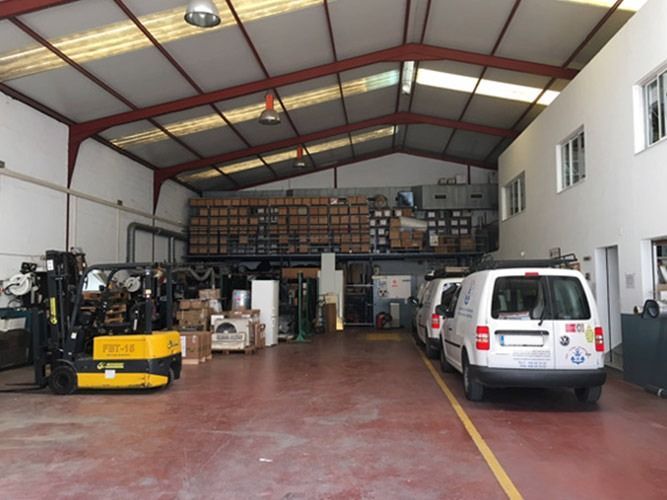 Dentro del almacén hay montacargas, furgonetas y estanterías con cajas. Línea amarilla en el suelo.