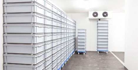 Contenedores de almacenamiento grises apilados en una sala de refrigeración industrial blanca, con una unidad de enfriamiento.