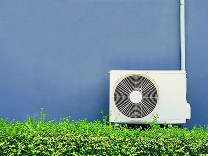 Unidad de aire acondicionado contra pared azul, con arbustos verdes.