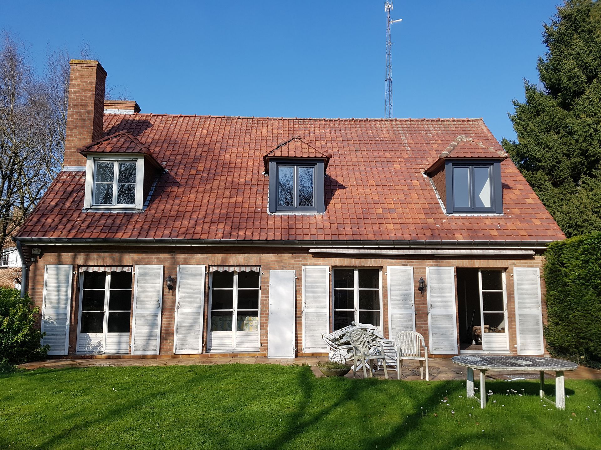 Maison en briques avec toit de tuiles rouges, volets blancs et pelouse verte.