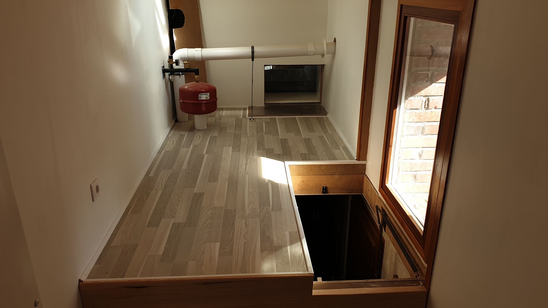 Une petite pièce avec un parquet, une fenêtre et un escalier descendant.