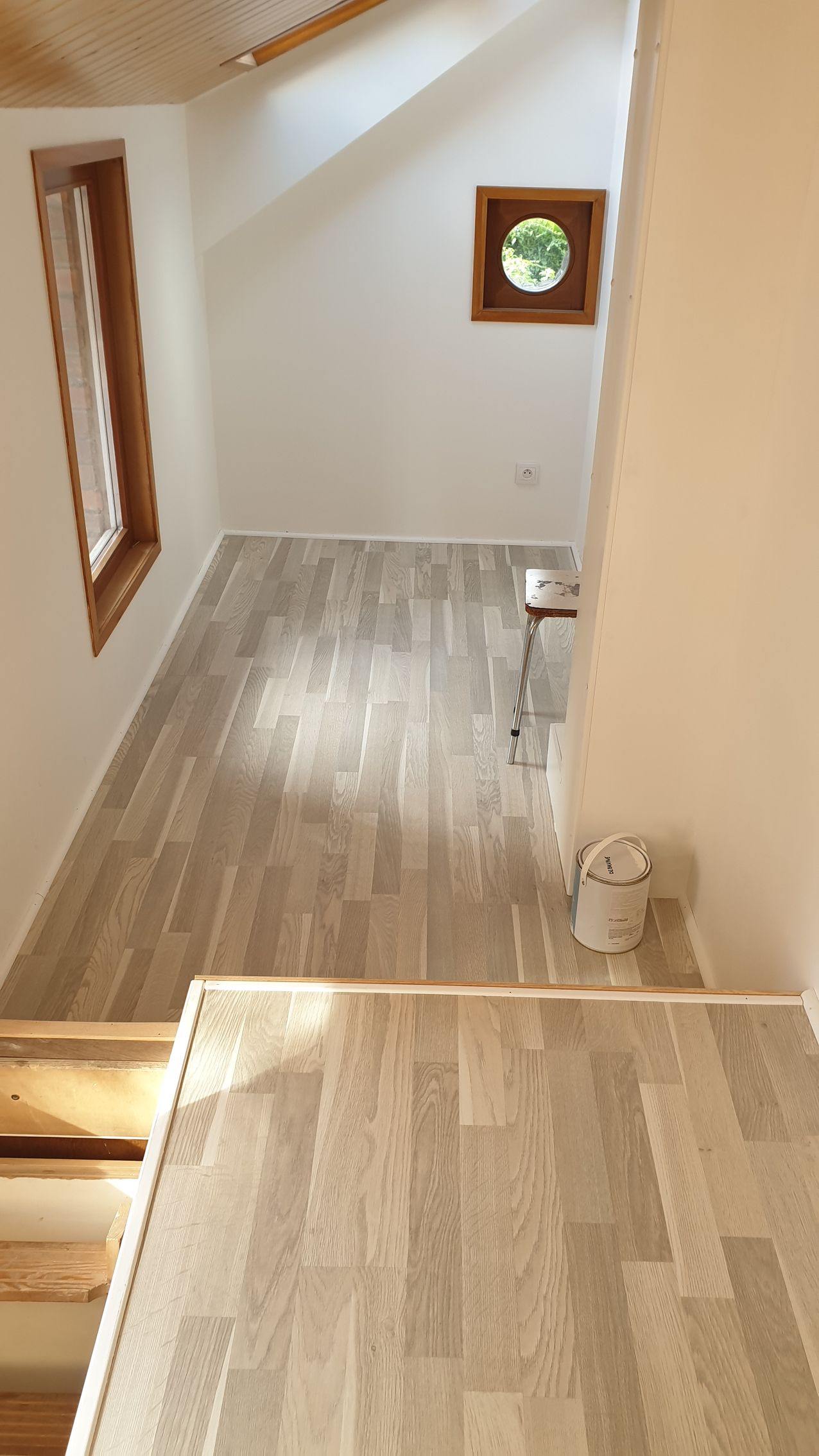 Vue intérieure d'une pièce avec parquet, une fenêtre et une fenêtre ronde. Un escalier descend dans la pièce.