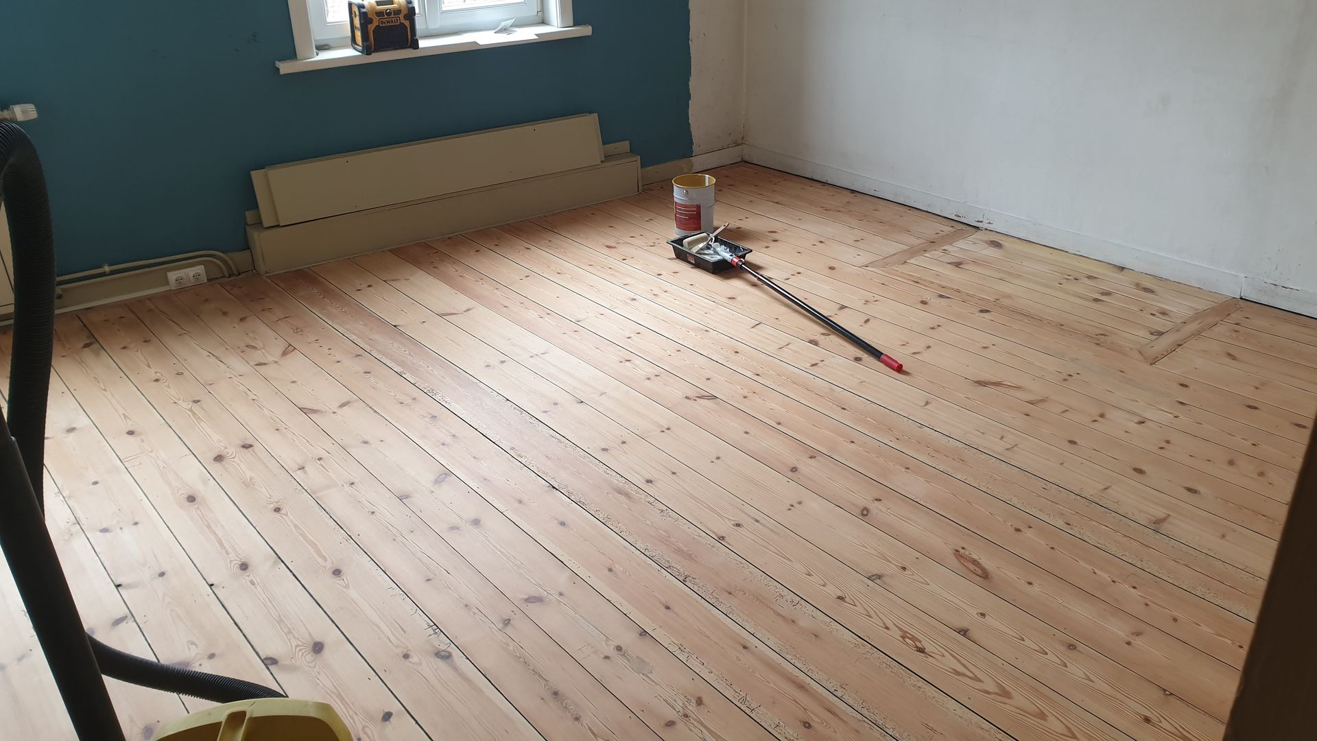 Un parquet est rénové à l'aide d'équipements de ponçage et de lames de bois dans une pièce aux murs bleus et blancs.