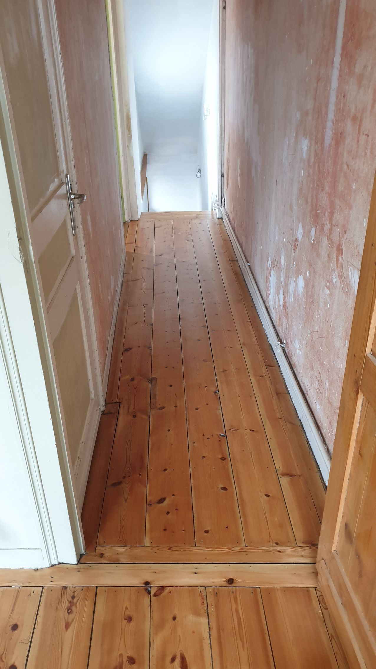 Couloir avec parquet, murs blancs, plafond mansardé et portes ouvertes. Un aspirateur et du matériel se trouvent dans le couloir.