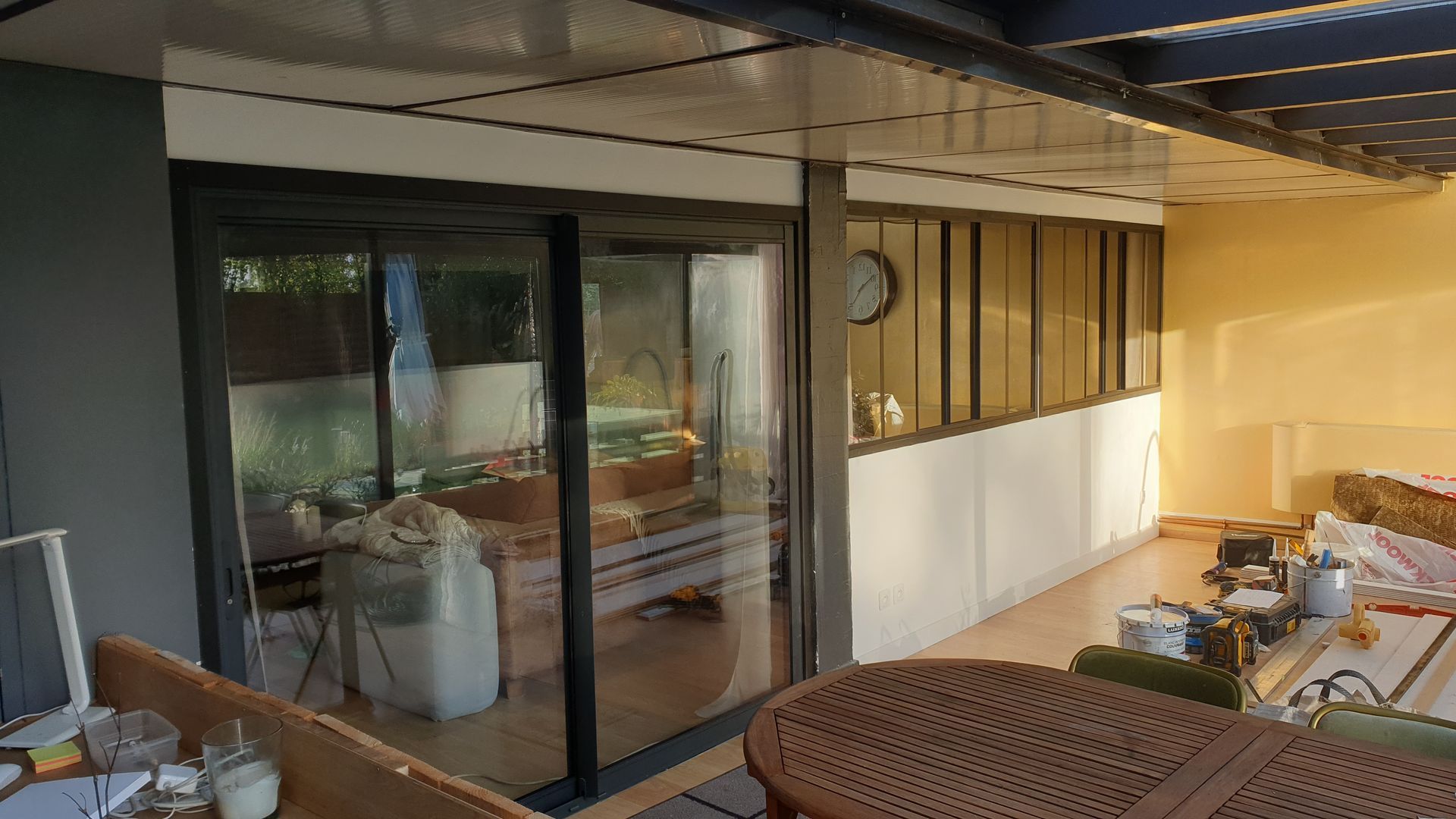 Une porte-fenêtre coulissante donne sur une terrasse. Mur blanc avec panneaux de verre et horloge. Espace extérieur avec quelques matériaux de construction.
