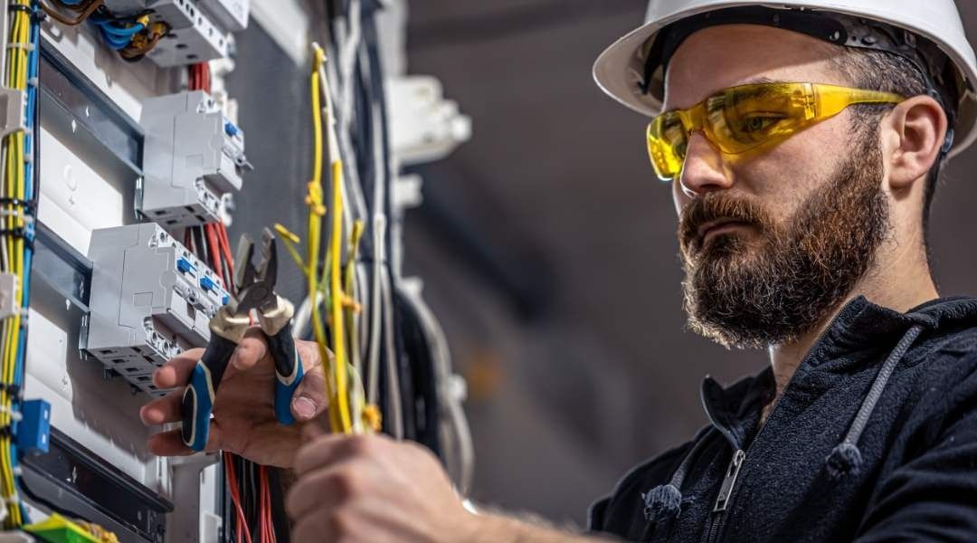 Un hombre que lleva casco y gafas de seguridad está trabajando en un panel eléctrico.