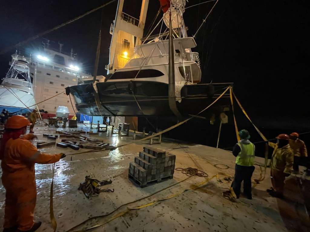 A tugboat is being lifted by a crane onto a larger vessel at night, workers oversee the operation.