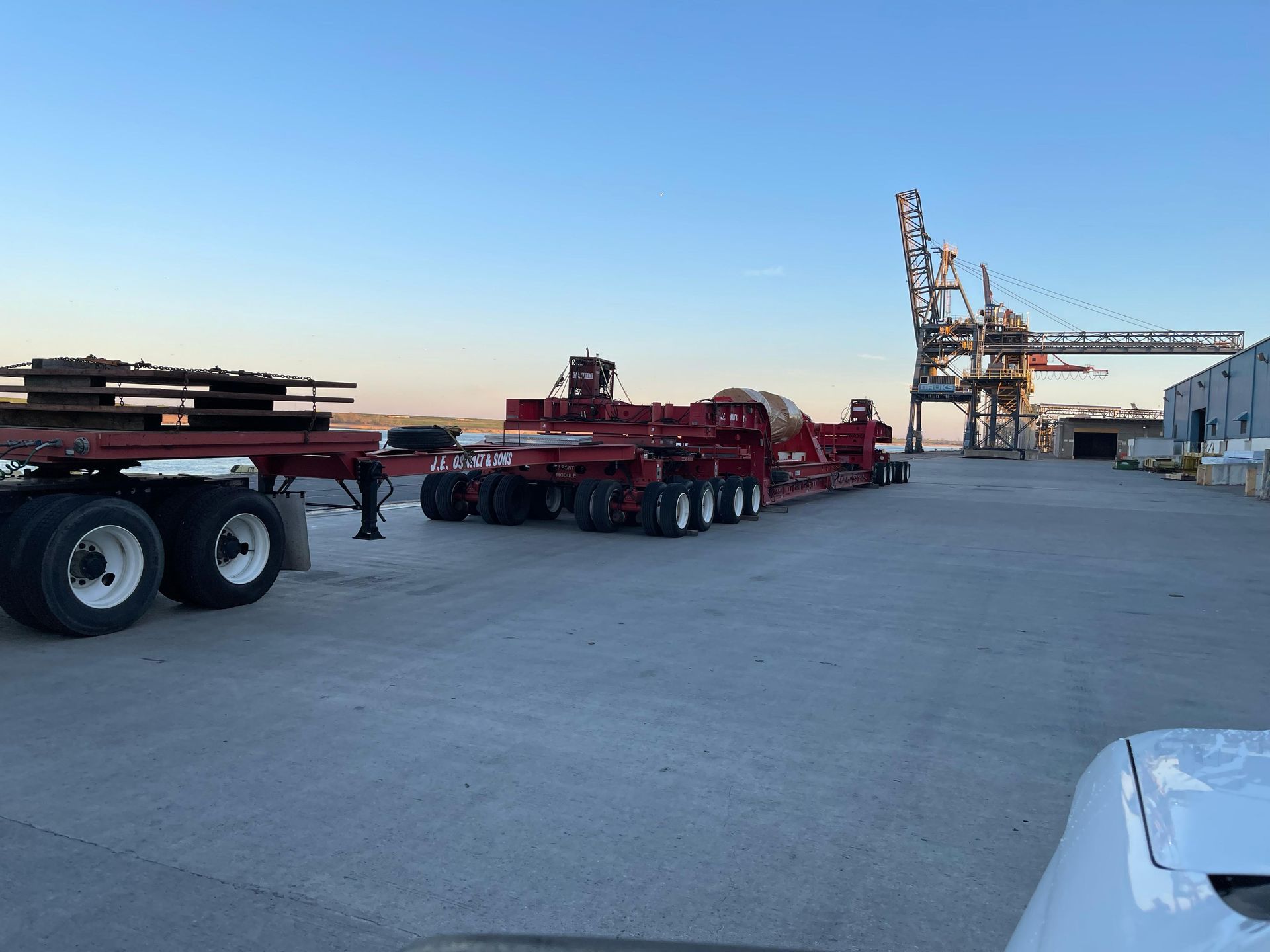 Red semi-truck trailers parked on a concrete dock near a large crane and buildings under a blue sky.