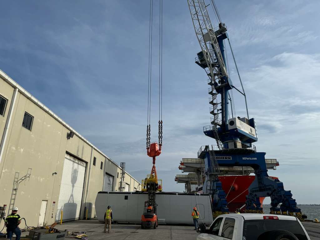 Crane lifting a container near a warehouse, workers nearby. Cloudy sky, industrial setting.