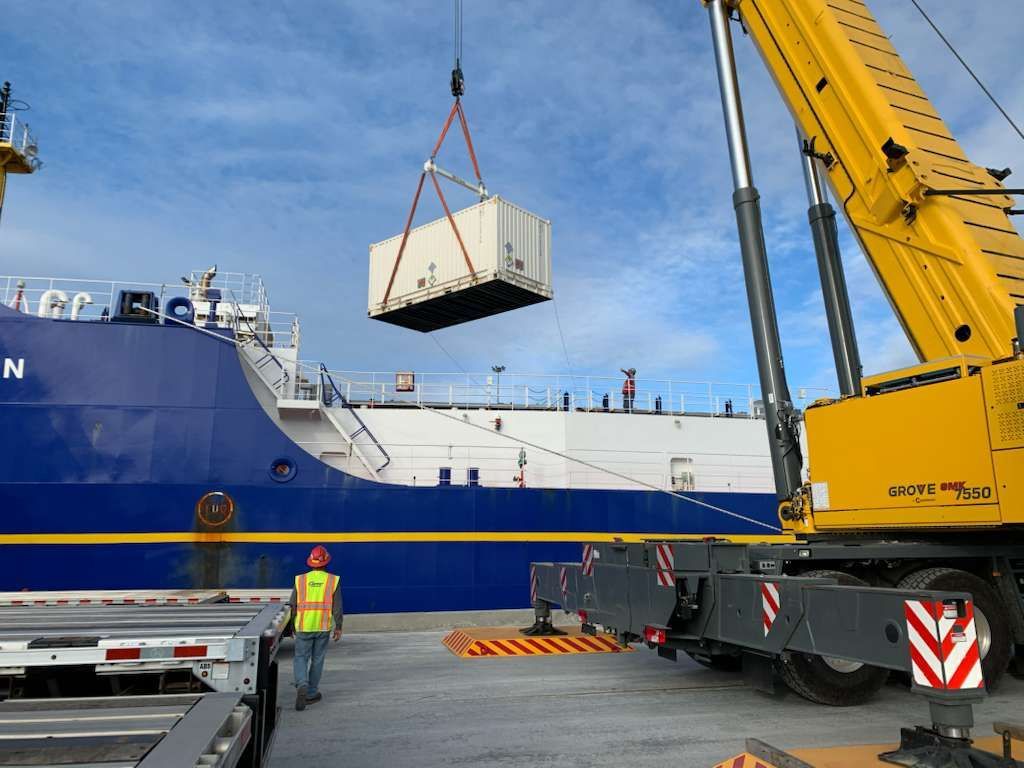 A crane lifting a white container from a dock next to a blue ship. A worker in a vest watches.